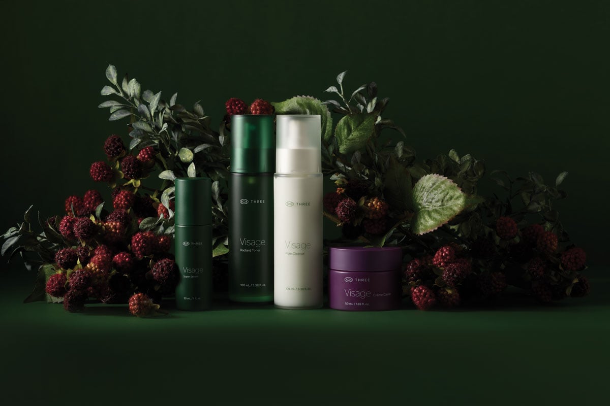 The complete Visage Skincare Collection by THREE displayed against a deep green botanical backdrop with berries and leaves, featuring Pure Cleanser, Radiant Toner, Super Serum, and Crème Caviar.