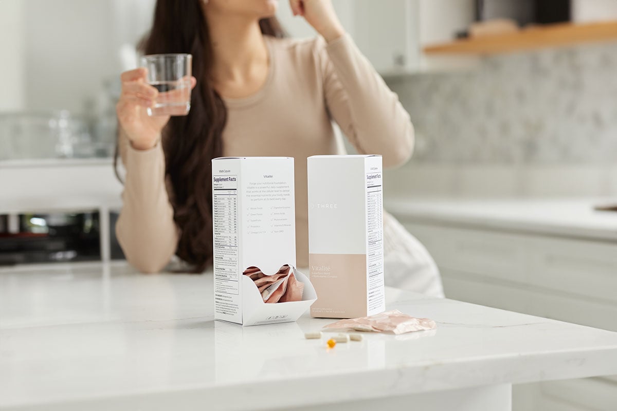 a woman in her kitchen taking a daily dose of multivitamin Vitalité by THREE