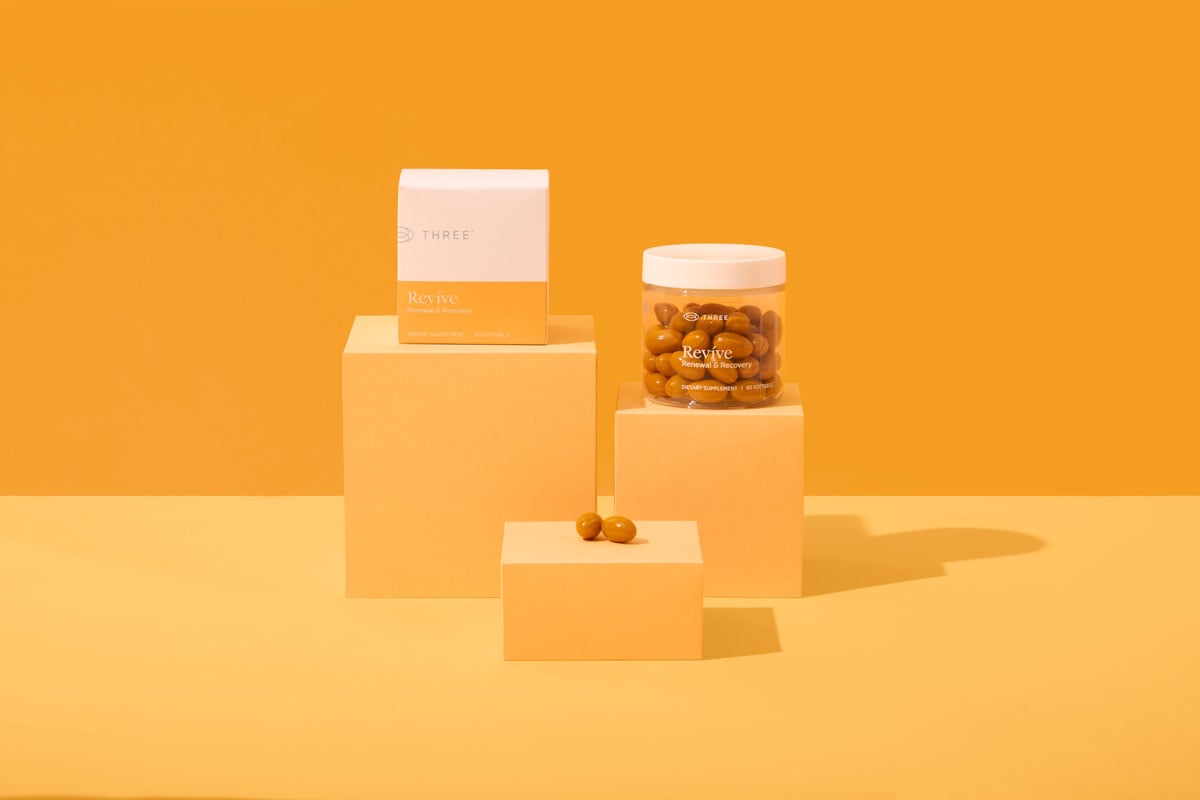 A clean, vibrant product display of Revíve by THREE, featuring a white and orange box, a clear jar filled with orange softgels, and additional capsules arranged on soft orange blocks against a matching orange background.